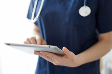 Doctor woman using tablet computer while standing near panorama window in clinic, close up. Physician or surgeon at work. Medicine concept