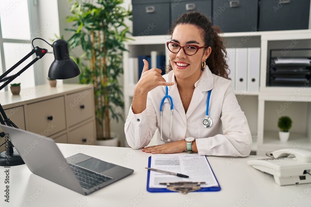 Sticker Young hispanic woman wearing doctor uniform and stethoscope smiling doing phone gesture with hand and fingers like talking on the telephone. communicating concepts.