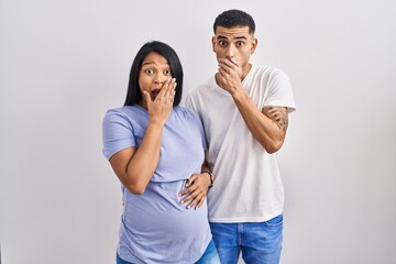 Naklejka premium Young hispanic couple expecting a baby standing over background shocked covering mouth with hands for mistake. secret concept.