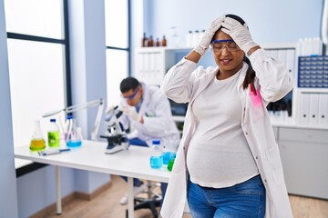 Obraz premium Young hispanic woman expecting a baby working at scientist laboratory suffering from headache desperate and stressed because pain and migraine. hands on head.