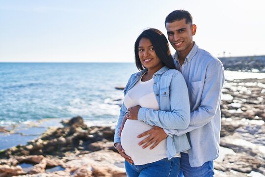 Young latin couple expecting baby hugging each other standing at seaside