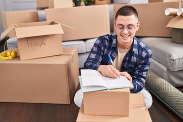 Fototapeta premium Young hispanic man writing on notebook sitting on floor at new home
