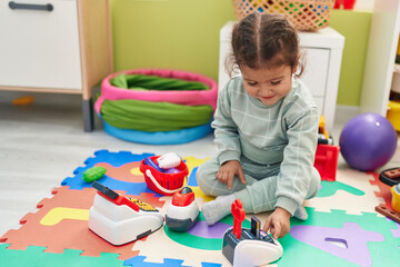 Adorable hispanic toddler playing with supermarket toy sitting on floor at kindergarten