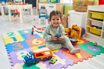 Adorable hispanic toddler playing with car toy sitting on floor at kindergarten