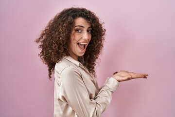 Hispanic woman with curly hair standing over pink background pointing aside with hands open palms showing copy space, presenting advertisement smiling excited happy