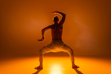 Fototapeta na wymiar back view of shirtless man practicing goddess yoga pose and gesturing on orange background.