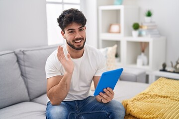 Hispanic man with beard using touchpad sitting on the sofa waiving saying hello happy and smiling,...