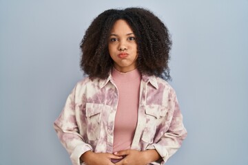 Young african american woman standing over blue background puffing cheeks with funny face. mouth...