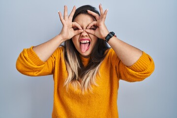 Young hispanic woman standing over isolated background doing ok gesture like binoculars sticking...