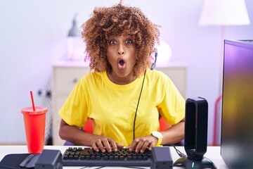 Young hispanic woman with curly hair playing video games wearing headphones afraid and shocked with surprise expression, fear and excited face.