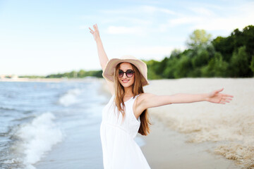 Fototapeta na wymiar Happy smiling woman in free happiness bliss on ocean beach standing with a hat, sunglasses, and raising hands. Portrait of a multicultural female model in white summer dress enjoying nature 