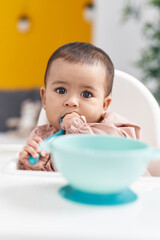 Adorable hispanic baby sitting on highchair sucking spoon at home