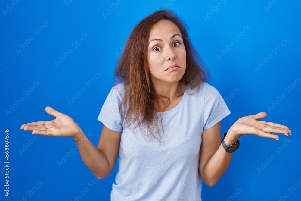 Poster brunette woman standing over blue background clueless and confused expression with arms and hands ra