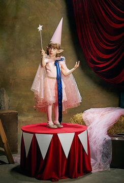 Emotional little kid, beautiful girl in image of fairy magical elf posing in cute pink dress over vintage circus background. Retro circus performance. Concept of dreams, art, vintage style, childhood