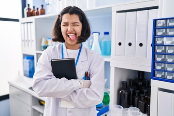 Young hispanic woman working at scientist laboratory sticking tongue out happy with funny expression.
