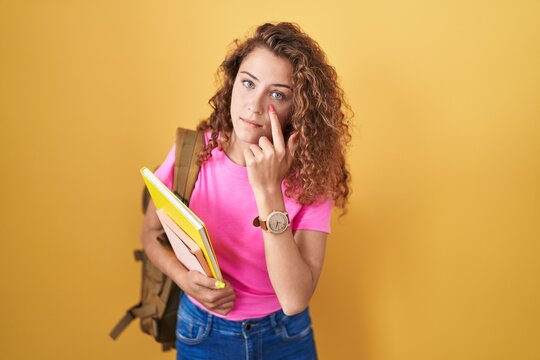 Young caucasian woman wearing student backpack and holding books pointing to the eye watching you gesture, suspicious expression