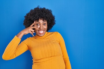 Fototapeta na wymiar Black woman with curly hair standing over blue background doing peace symbol with fingers over face, smiling cheerful showing victory