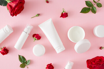 Organic skincare concept. Flat lay composition made of cosmetic bottles without label, cream jars and red flowers with leaves on pastel pink background. Cosmetics mockup idea.