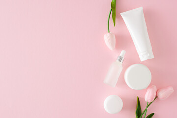 Organic skincare concept. Flat lay photo of cosmetic tubes without label, cream jars, dropper bottle and spring flowers on pastel pink background with empty space. Cosmetics mockup idea.