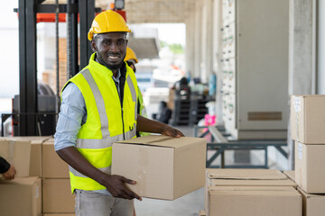 Black Man worker in safety vest and yellow helmet holding cardboard box preparing product for shipment at warehouse factory