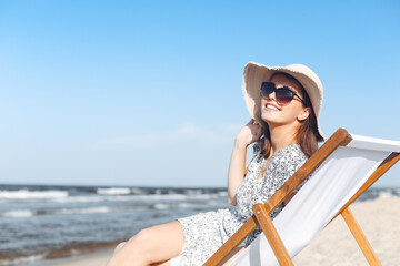 Happy brunette woman wearing sunglasses and hat relaxing on a wooden deck chair at the ocean beach