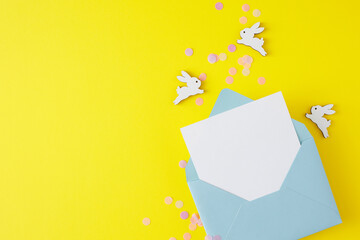 Easter card concept. Flat lay photo of open envelope with white card, confetti and cute easter bunnies on yellow background with empty space. Holiday invitation card idea.