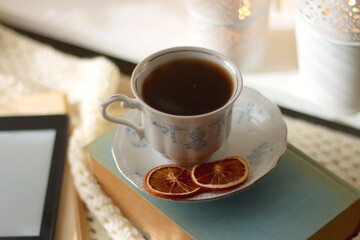 Vintage books, reading glasses, e-reader, cup of tea, bowl of biscuits and lit candle on the table. Selective focus.