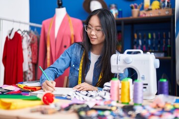 Young chinese woman tailor smiling confident drawing on notebook at clothing factory