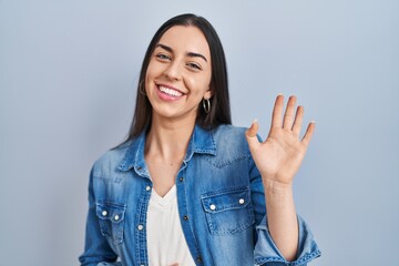 Hispanic woman standing over blue background waiving saying hello happy and smiling, friendly...