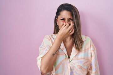 Blonde woman standing over pink background smelling something stinky and disgusting, intolerable...