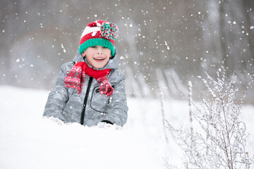 Child in winter for a walk. A little boy, in a red knitted hat, sits in a snowdrift.