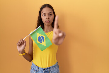 Young hispanic woman holding brazil flag pointing with finger up and angry expression, showing no gesture