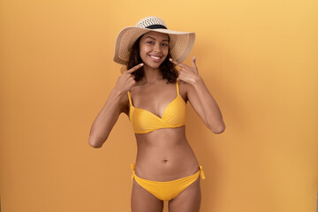 Young hispanic woman wearing bikini and summer hat smiling cheerful showing and pointing with fingers teeth and mouth. dental health concept.