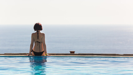 A woman in a black swimsuit is sitting with her back turned on the edge of the pool.