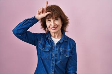 Middle age woman standing over pink background making fun of people with fingers on forehead doing...