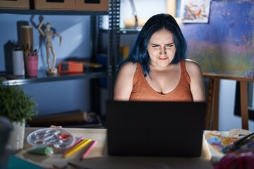 Fototapeta na wymiar Young modern girl with blue hair sitting at art studio with laptop at night with hand on stomach because nausea, painful disease feeling unwell. ache concept.