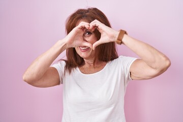 Middle age woman standing over pink background doing heart shape with hand and fingers smiling looking through sign