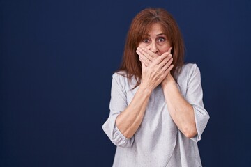 Middle age woman standing over blue background shocked covering mouth with hands for mistake....