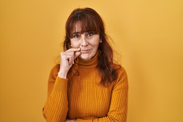 Middle age hispanic woman standing over yellow background mouth and lips shut as zip with fingers. secret and silent, taboo talking