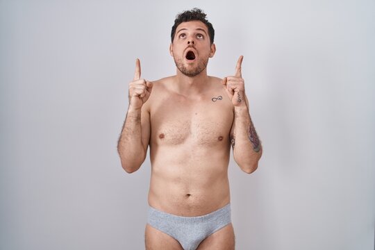 Young hispanic man standing shirtless wearing underware amazed and surprised looking up and pointing with fingers and raised arms.