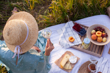 A woman in a blue dress and straw hat with short hair sitting on a white blanket holding glass and drinking white wine. Concept of having picnic in a city park during summer holidays or weekends. 