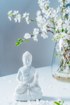 Decorative white Buddha statuette with blooming tree branches in the vase on the light background. Meditation and relaxation ritual. Buddha birthday. Vertical card. Selective focus