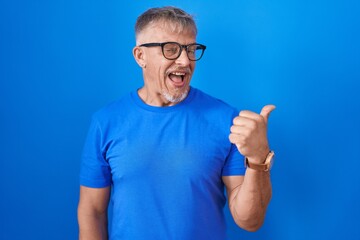 Hispanic man with grey hair standing over blue background smiling with happy face looking and pointing to the side with thumb up.