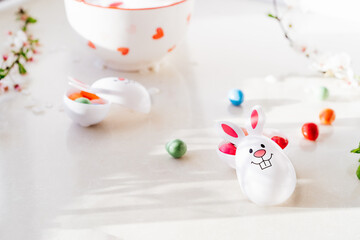 Obraz na płótnie Canvas Close up Funny Easter eggs containers with bunny ears filled with candy and chocolate on the white kitchen table with blooming branches. Egg hunt. Festival spring Easter composition. Selective focus