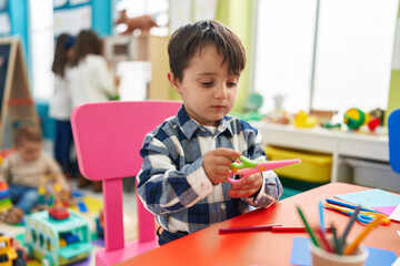 Adorable hispanic boy student holding scissors at kindergarten