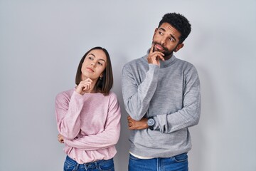 Young hispanic couple standing together with hand on chin thinking about question, pensive expression. smiling with thoughtful face. doubt concept.