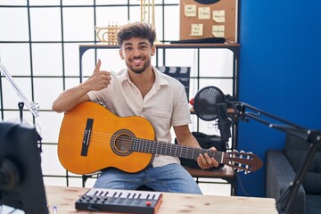 Arab man with beard playing classic guitar at music studio smiling happy and positive, thumb up doing excellent and approval sign