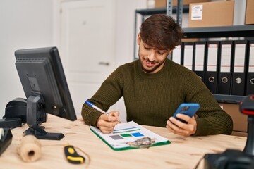 Young arab man ecommerce business worker writing on document using smartphone at office