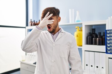 Arab man with beard working at scientist laboratory peeking in shock covering face and eyes with...