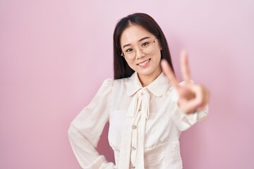 Young chinese woman standing over pink background smiling looking to the camera showing fingers doing victory sign. number two.